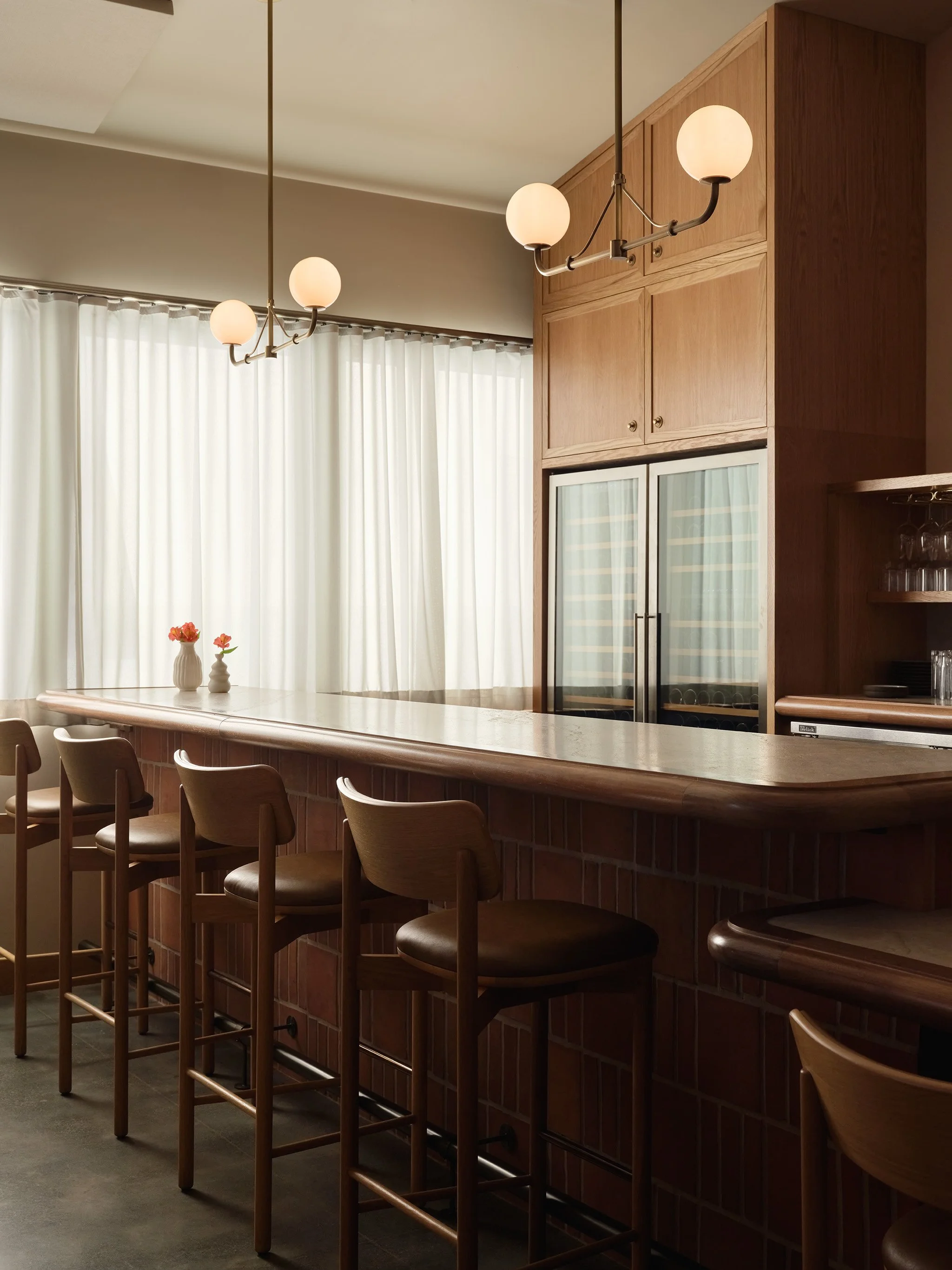 Interior of a bar or kitchen area with a wooden counter, bar stools, built-in wooden cabinets, a wine cooler, and hanging globe pendant lights. There are white curtains and flower vases on the counter.