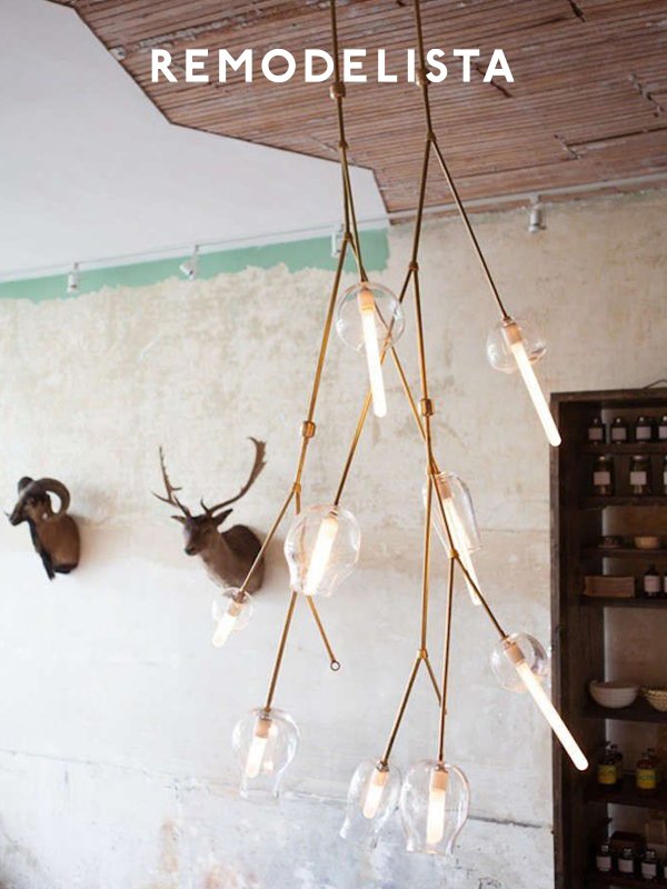 Modern hanging light fixture with multiple glass bulbs and gold cords in a rustic interior. Two mounted antelope heads on the wall and a wooden shelf with jars are visible.