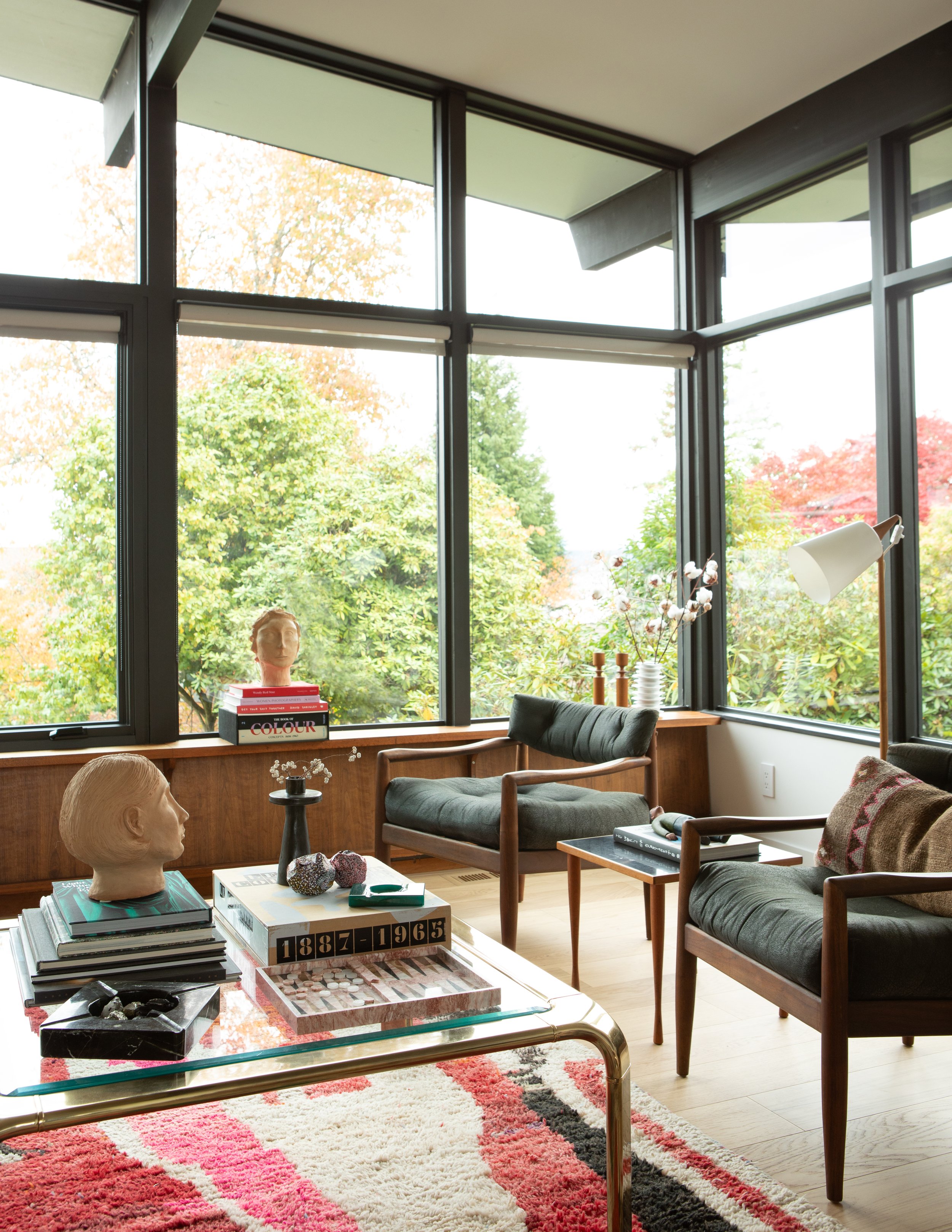 Interior view of a modern living room with large corner windows overlooking trees with colorful fall foliage. The room features a wooden side table with a decorative bust, books, and rocks, a colorful rug, two armchairs with dark cushions, and a glas