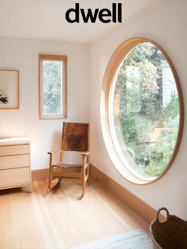 A room with a large circular window showing greenery outside, a wooden chair, a small wooden dresser, and a framed picture on the wall.