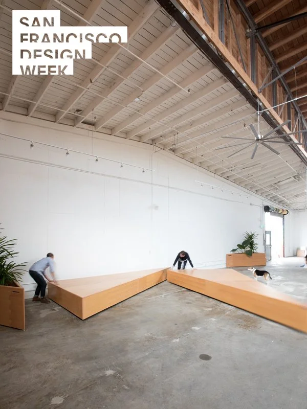 People constructing a wooden seating area inside an industrial-style space at San Francisco Design Week, with potted plants and large open space.