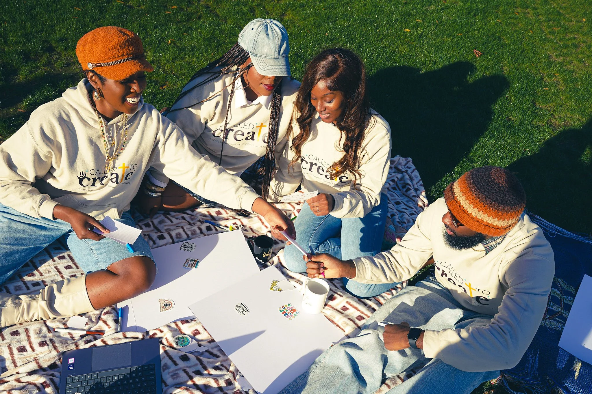 Group of four women sitting on a blanket on the grass, working together with papers, stickers, and laptop, all wearing matching white shirts with a logo and text.
