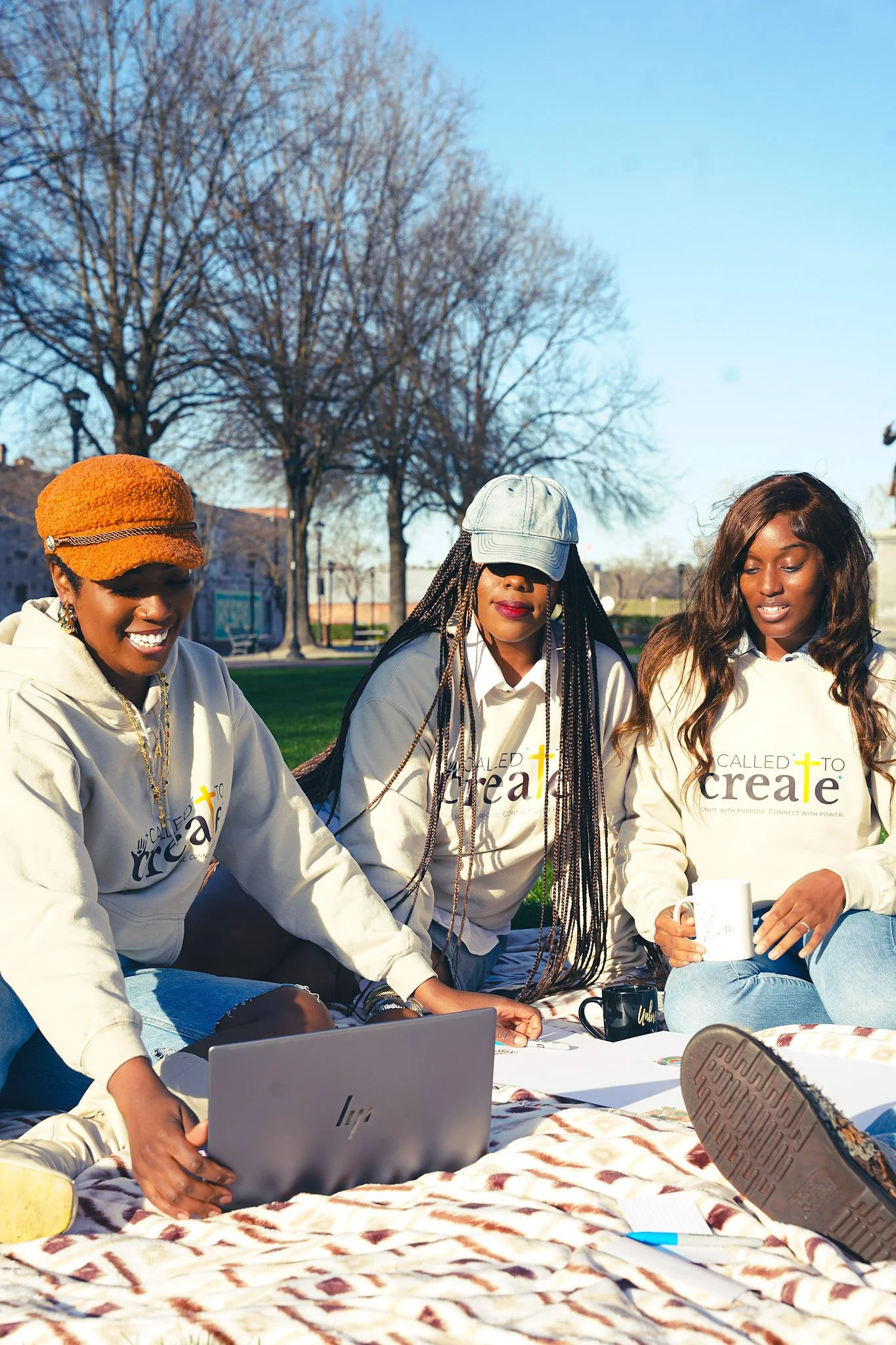 Three women sitting outdoors on a blanket, engaging with laptops, coffee mugs, and a notebook, in a park during daytime with bare trees and clear blue sky.