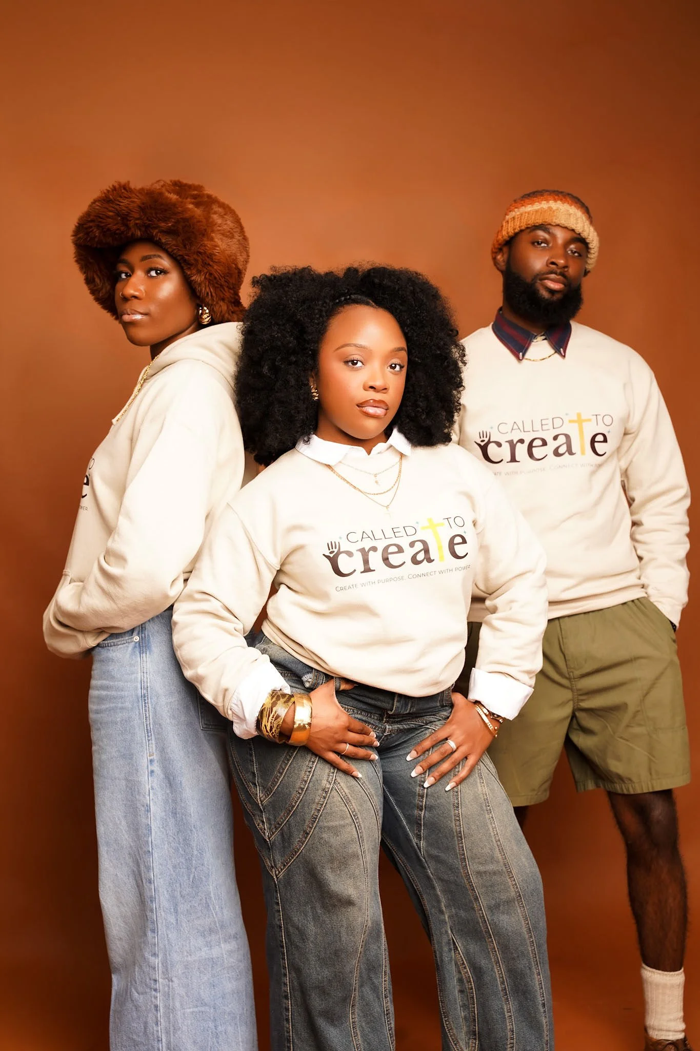 Three young adults standing together against a brown background, wearing beige sweatshirts with the word 'create' and a cross symbol, dressed casually with unique accessories.