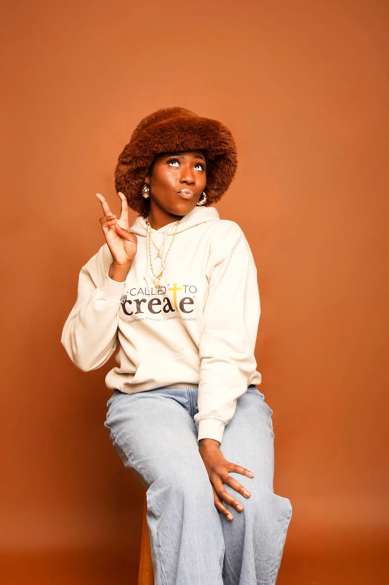 A woman wearing a large brown fur hat, earrings, and a cream hoodie with the words 'called to create' printed on it, sitting against a plain brown background.