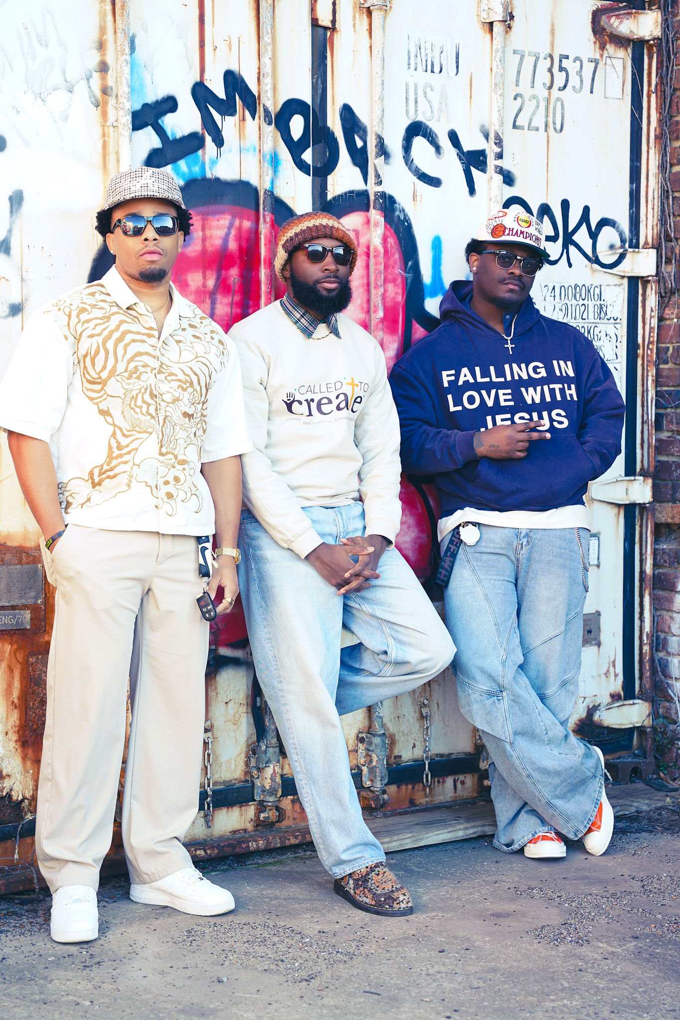Three young men standing in front of a weathered, graffiti-covered metal container. They are wearing casual streetwear, including hoodies, jeans, and sneakers, each with sunglasses and unique hats or headwear.