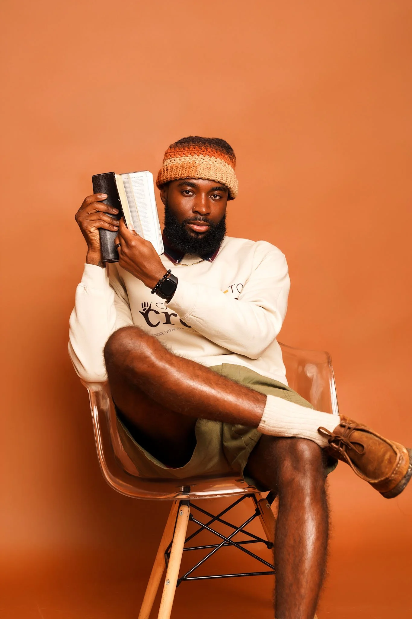 A man sitting on a transparent chair against a orange background, wearing a beanie, a white long-sleeve shirt, khaki shorts, and brown shoes, holding a black wallet and a book, with one leg bent and the other extended.