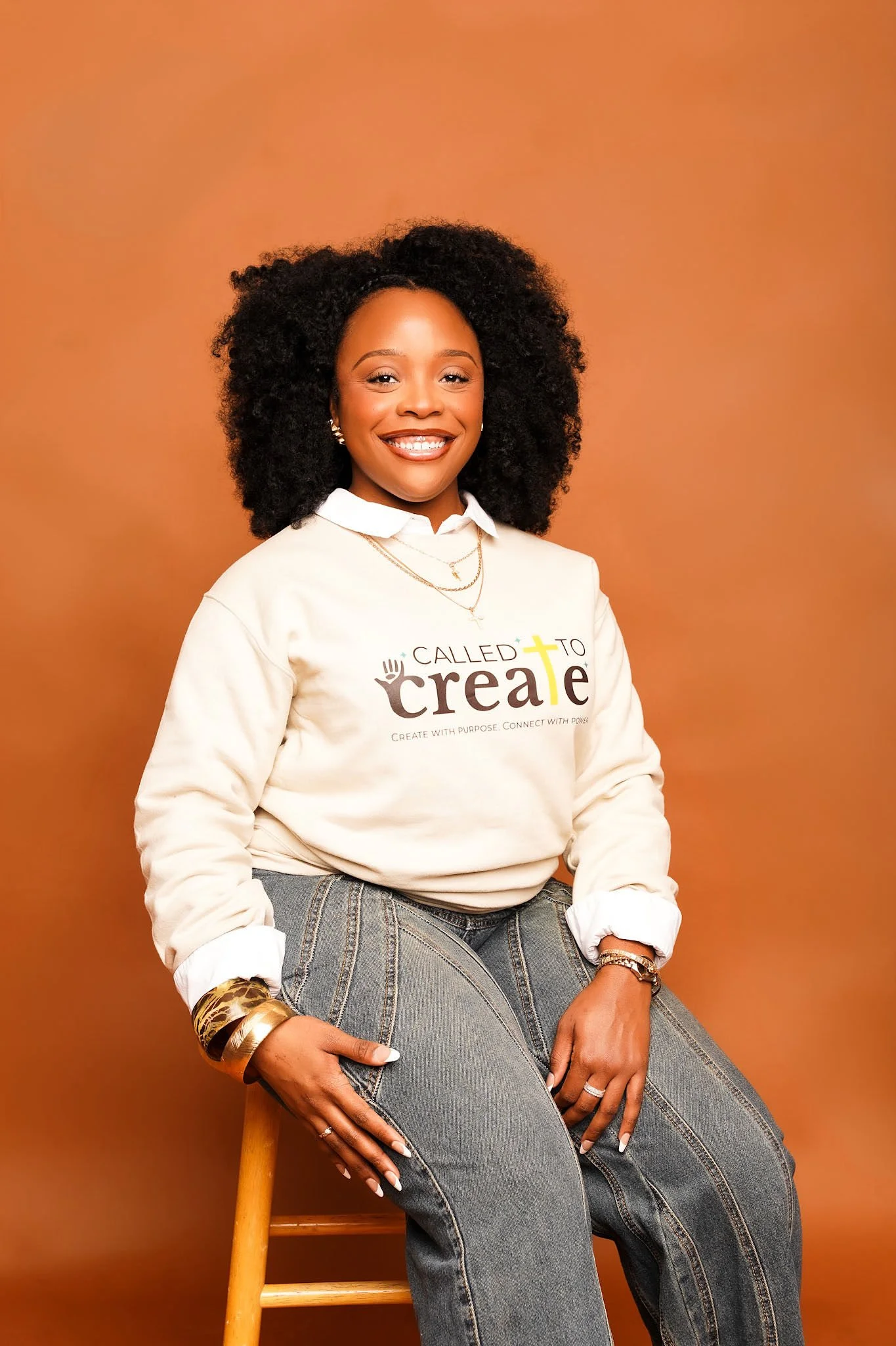 A woman with curly black hair wearing a white sweatshirt with the word 'Create' and a cross in yellow, gold hoop earrings, and grey jeans, sitting on a wooden chair against a warm orange background, smiling and looking at the camera.