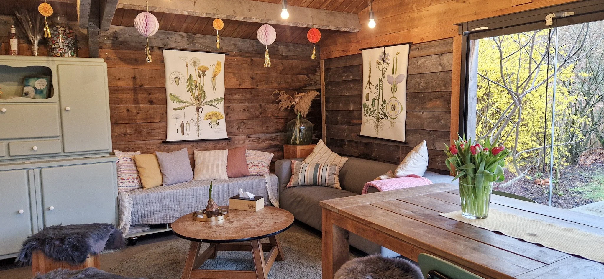 A cozy living room with rustic wooden walls, decorated with botanical prints, string lights, and hanging paper lanterns. There is a beige sofa with pillows, a round wooden coffee table, and a vase of pink tulips on a wooden dining table near a large window showing greenery outside.