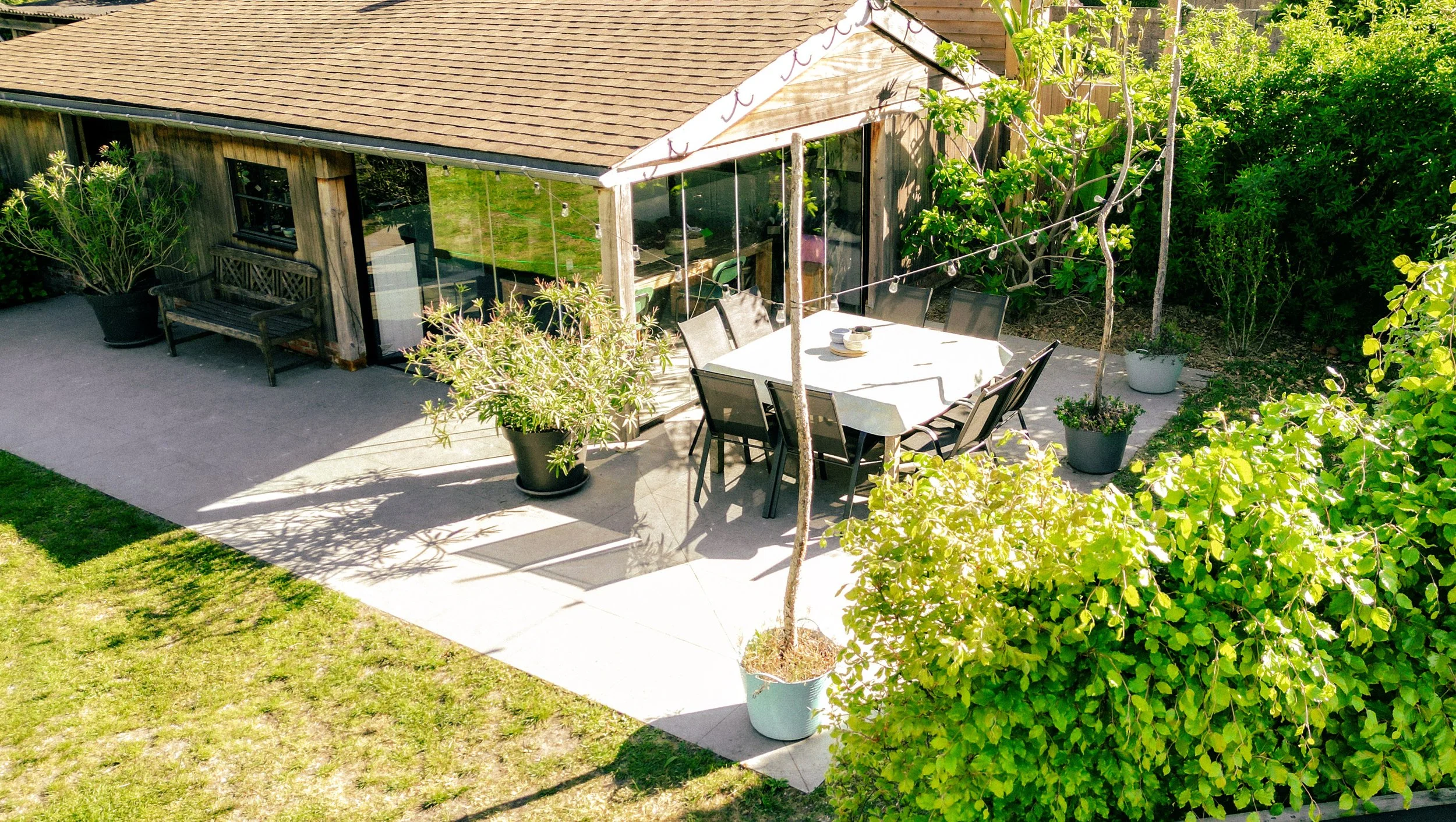 A backyard patio with a dining table and chairs, potted plants, and a wooden house with large windows, surrounded by greenery.