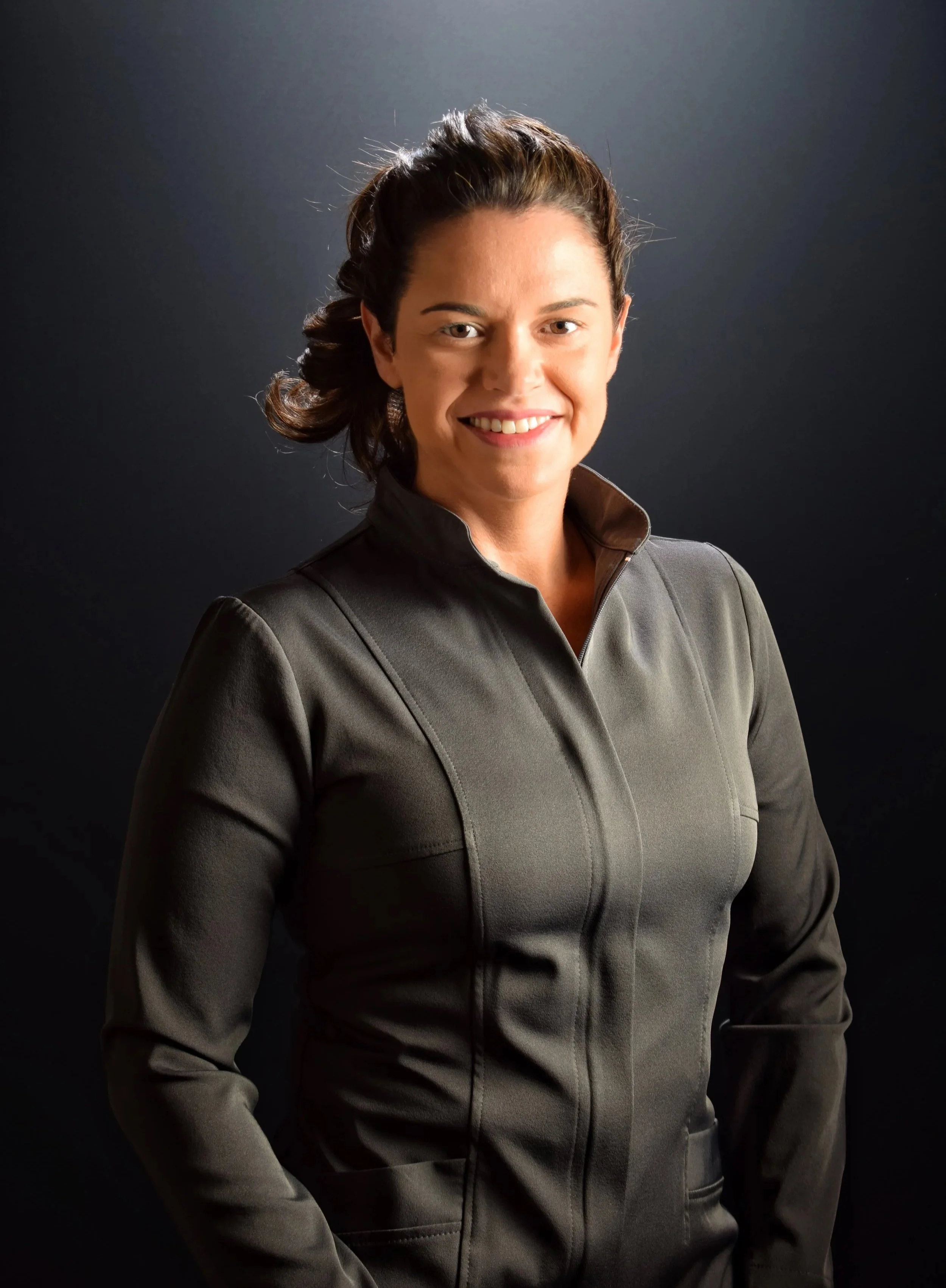 Mulher jovem sorrindo, vestindo jaqueta cinza, com cabelo castanho escuro preso em um coque, em fundo preto.