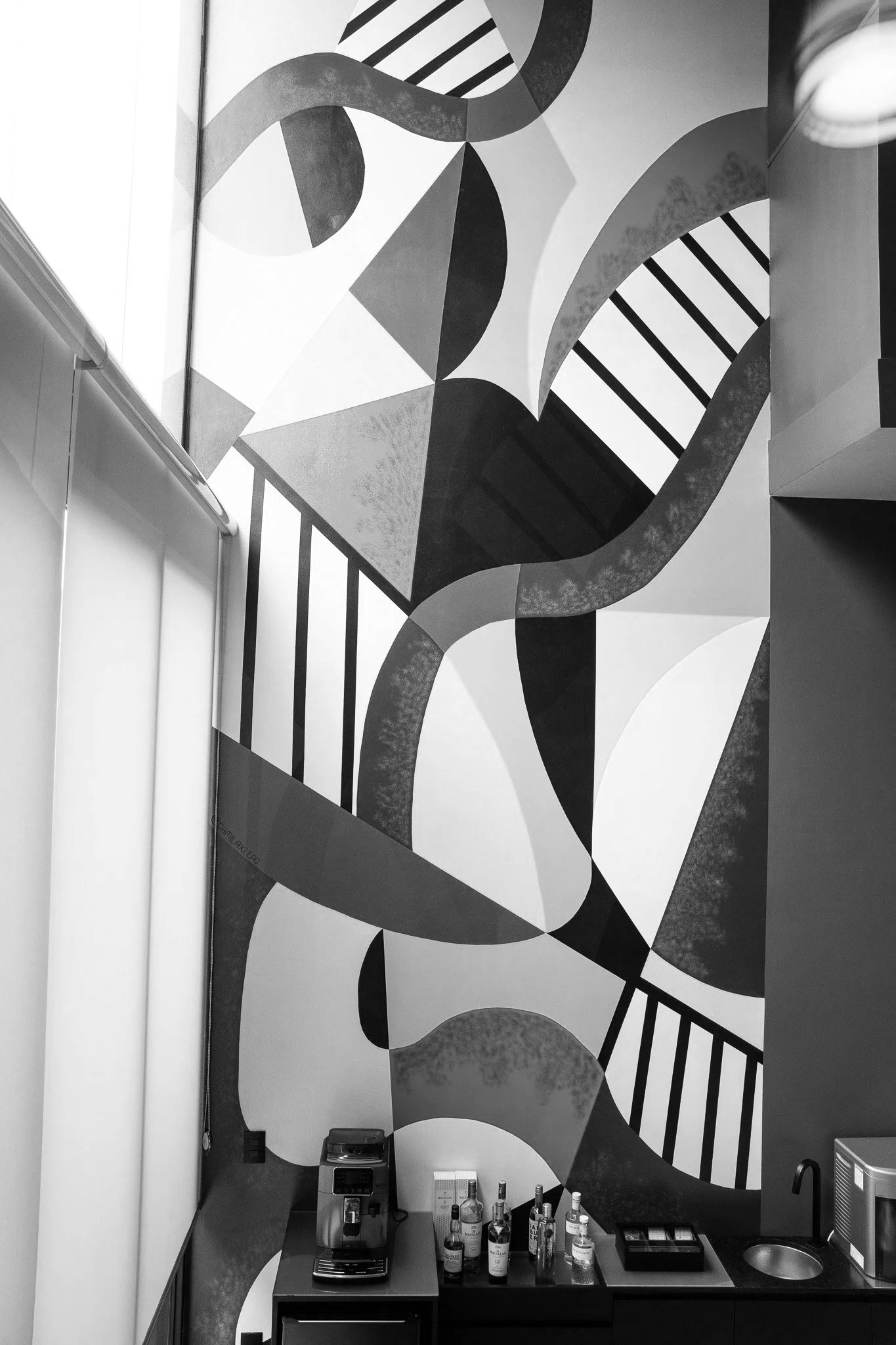 Parede com arte abstrata em tons de preto, branco e cinza na área de um bar ou kitchenette, com bebidas e equipamentos na bancada.