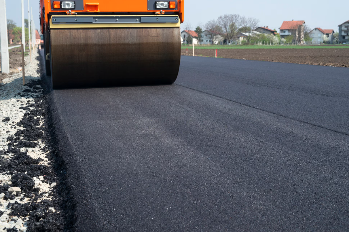 A road roller compacting freshly laid asphalt on a road under construction in a rural area with houses and trees in the background.