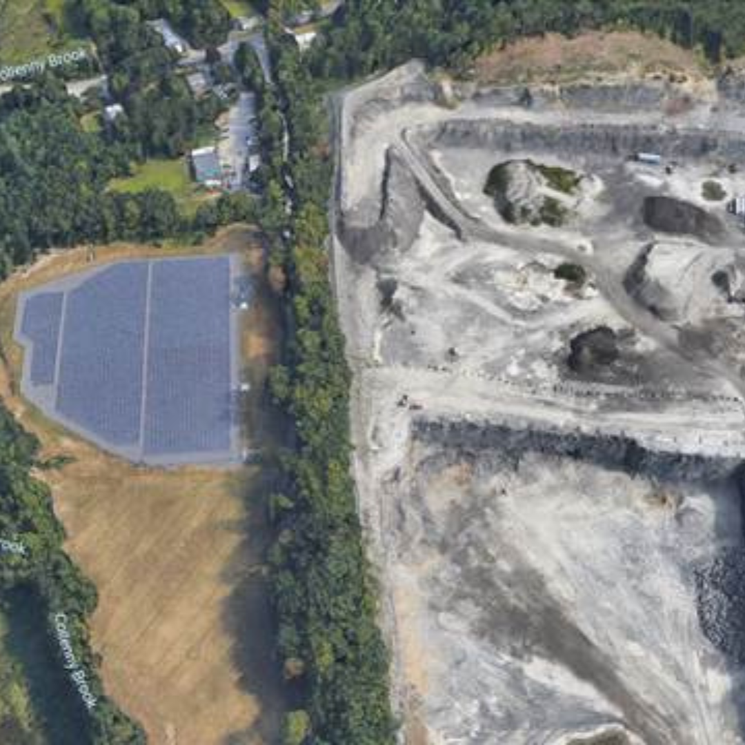 Aerial view of a large bare excavation site with rocks and dirt, next to a parking lot and a green area with trees.