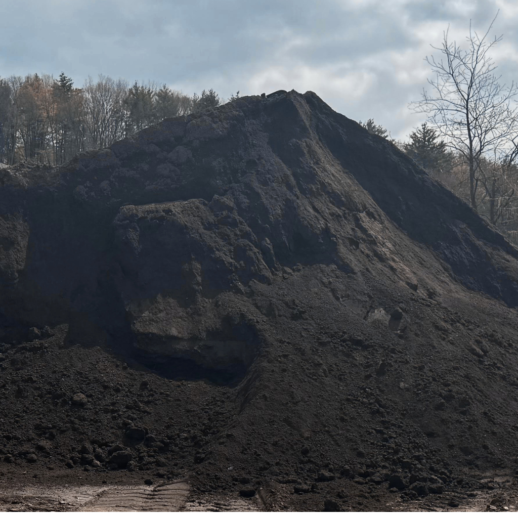 A large pile of black volcanic ash and rock with surrounding trees under a cloudy sky.