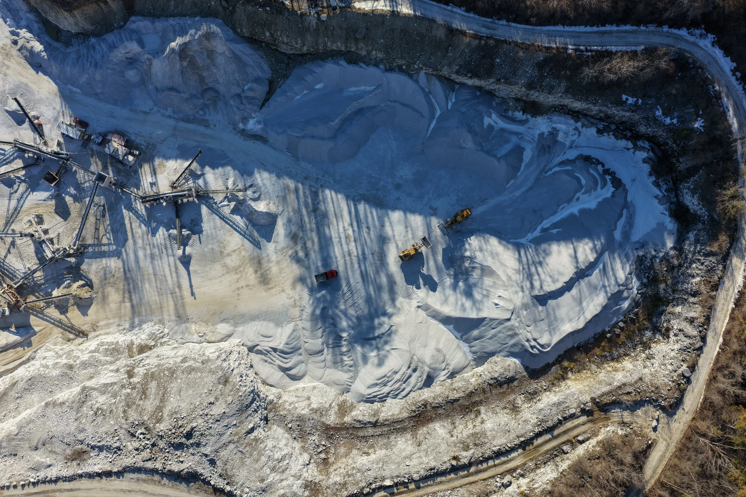 An aerial view of a winter oil extraction operation at a frozen lake, with pipelines, equipment, and vehicles on snow-covered ice surrounded by rocky cliffs.