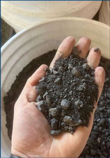 A hand holding black charcoal or crushed coal, with a large container of similar material in the background.