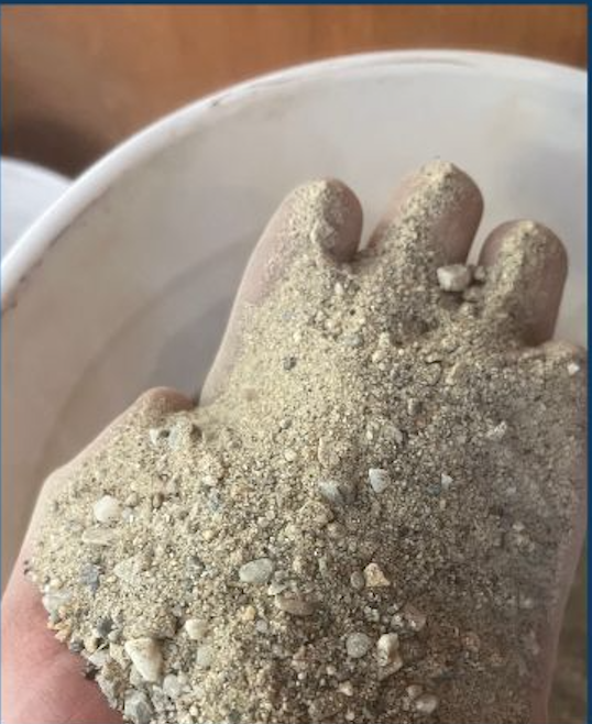 A person wearing a glove is holding a handful of gravel or sand with small rocks in front of a white container or basin.