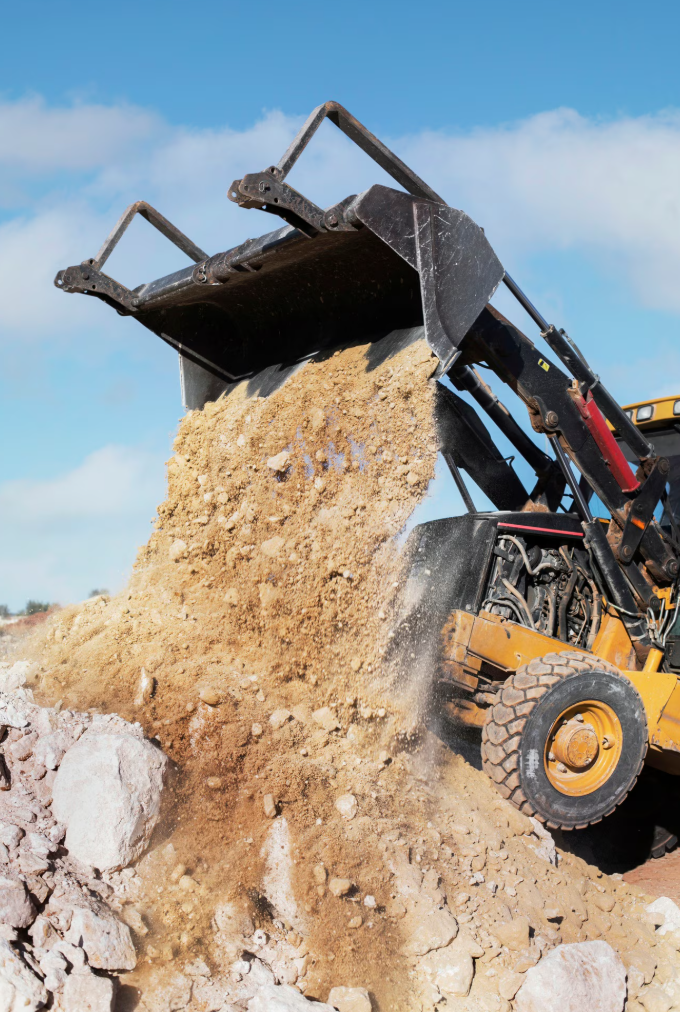 A front loader construction vehicle dumping dirt and rocks at a work site under a blue sky.