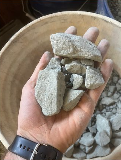 A person's hand holding several irregularly shaped rocks over a round container filled with more rocks.