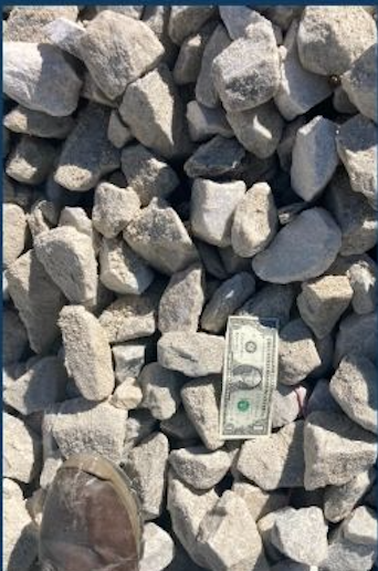 Pile of gray rocks with a dollar bill and a person's foot in a shoe for scale.