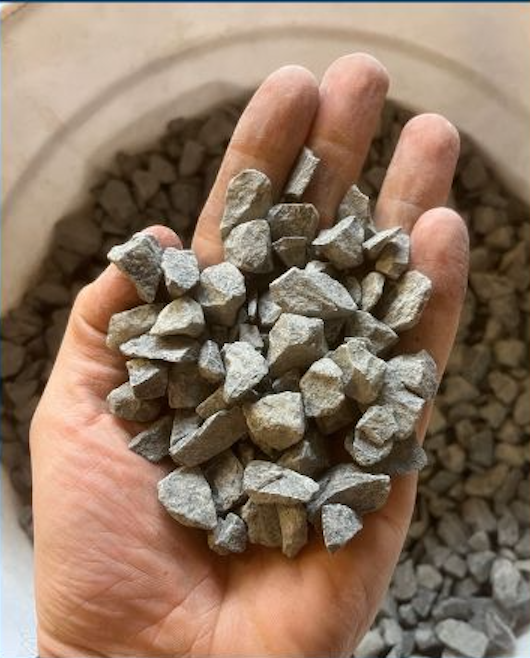A hand holding small gray crushed stone gravel, with a background of more gravel in a container.