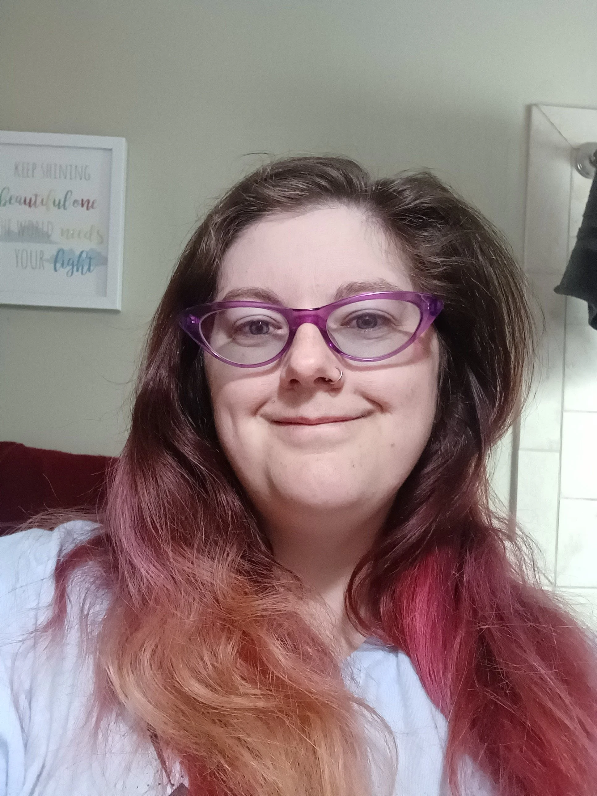 A woman with glasses and a nose ring smiling in a room with a framed quote on the wall.