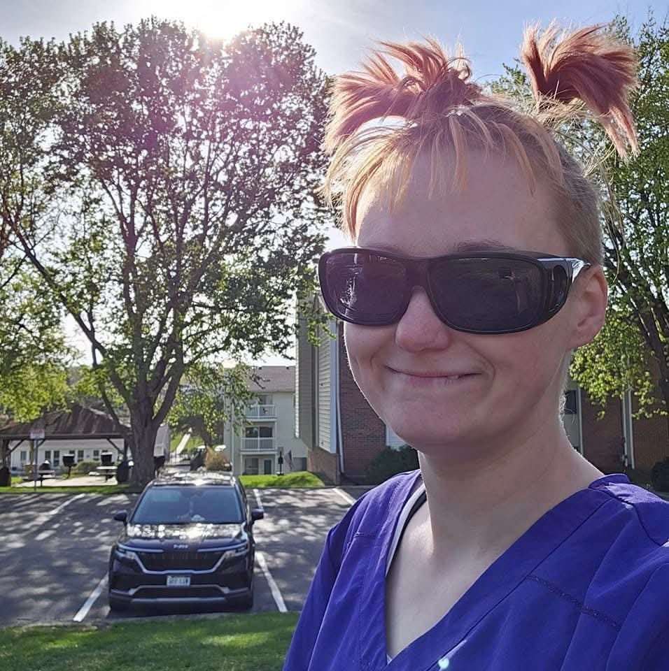 Person with short reddish hair styled in two buns, wearing large black sunglasses and blue scrubs, smiling outdoors with trees and parked cars in the background.