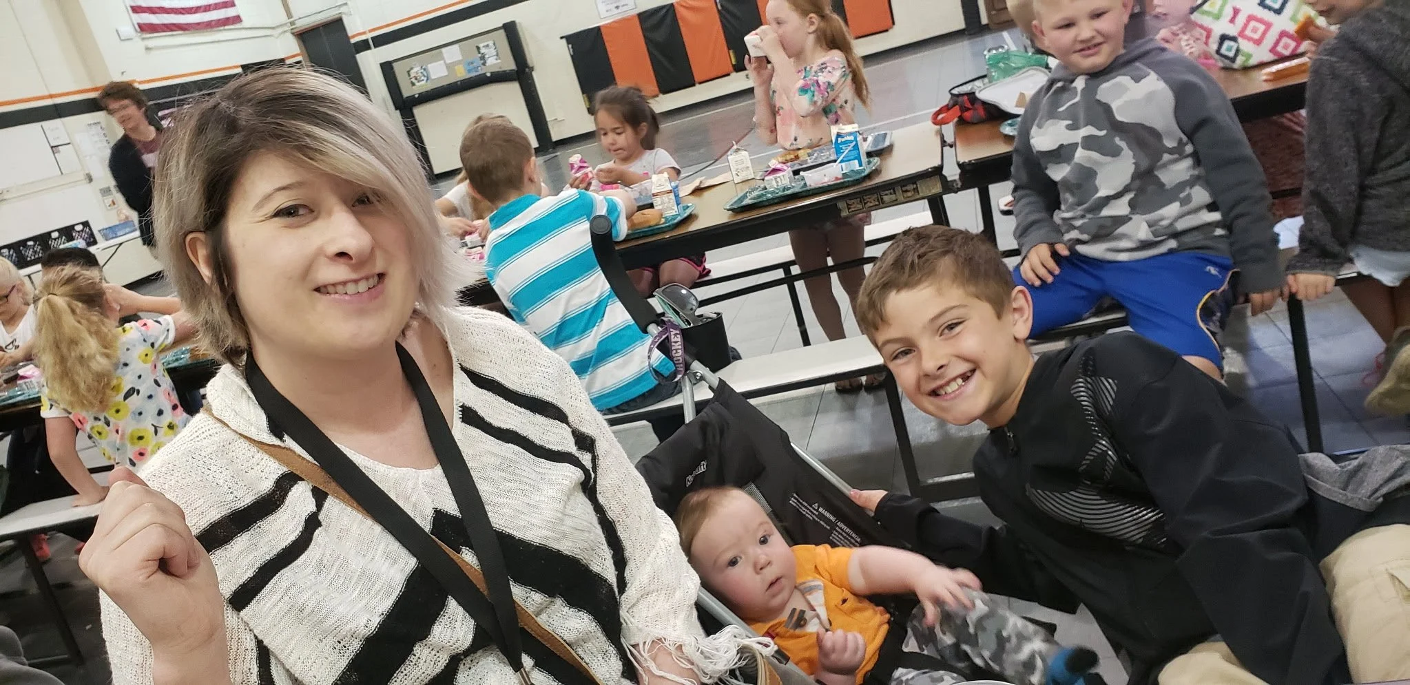 Group of children and a woman sitting at a cafeteria table enjoying a meal together with smiling faces.