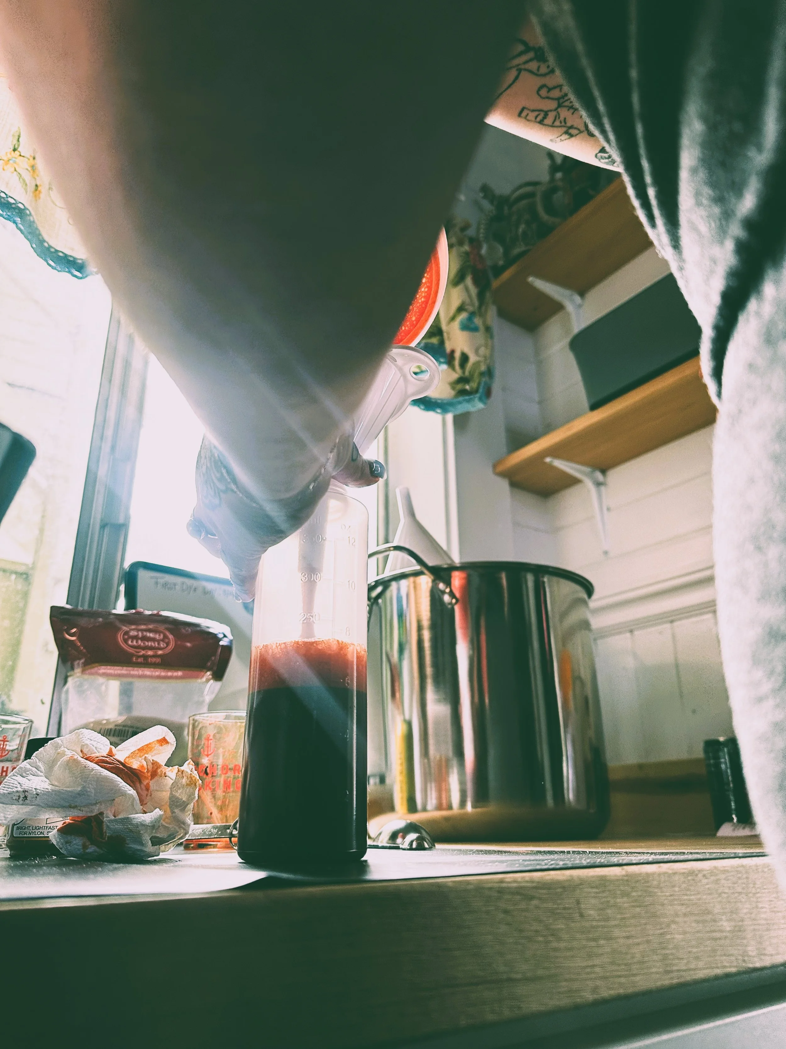 Inside a kitchen, taken from a low angle view, showing a black coffee mug on a wooden surface, with a reflection of a window in the background and various kitchen items such as a rice cooker, dish towel, and bottles on the counter and shelves.