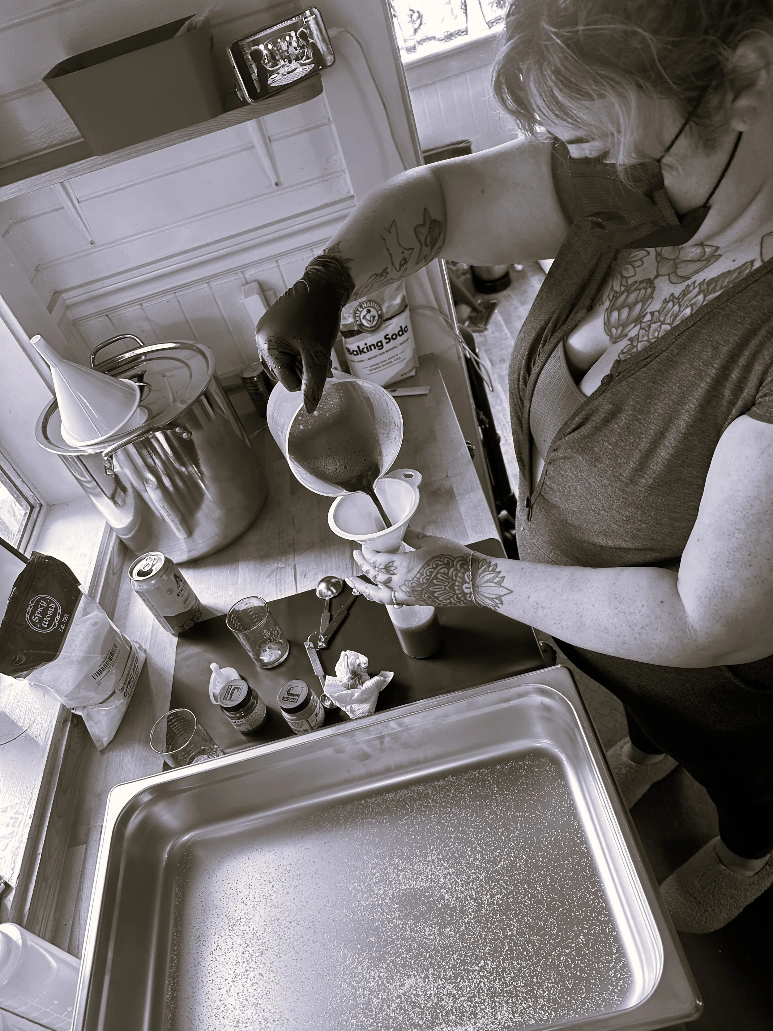 A person with tattoos on their arm, wearing a face mask and gloves, pours liquid from a pitcher into a cup in a kitchen. The kitchen counter has various items including spice jars and a can, and a large stainless steel container is on the counter. A small television on the wall displays a cooking show.