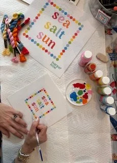 Craft project with a paper that says 'sea salt sun' in colorful letters, art supplies including paints, brushes, and a palette, and a woven keychain or tassel on a white table.