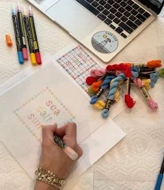 Hand writing on a square paper with colorful pens, surrounded by a laptop, a mouse, and a collection of colorful thread skeins on a white desk.