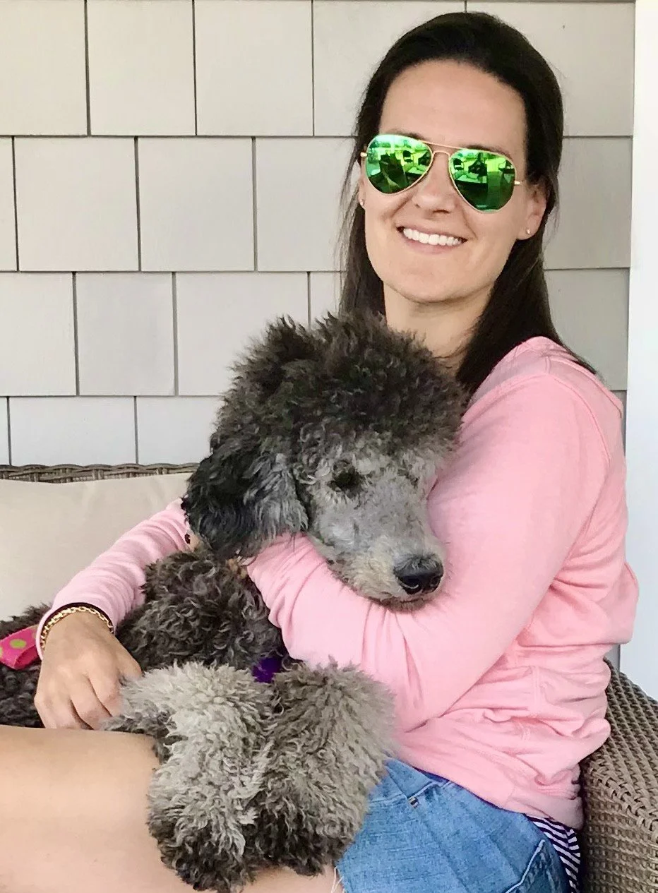 A woman wearing green sunglasses and a pink long-sleeve shirt sitting on a porch, hugging a curly-coated gray dog with a fluffy black face.