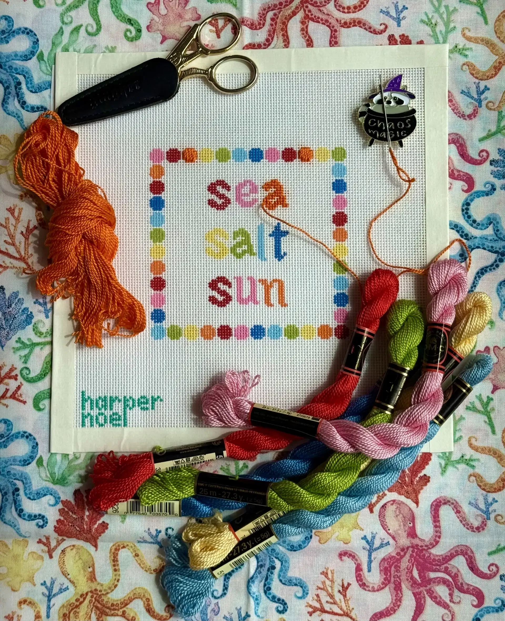 Colorful embroidery floss arranged in a circle on a cross-stitch fabric with the words "sea, salt, sun" stitched in colorful letters. A pair of small scissors and embroidery floss are also on the fabric, all on a multicolored, coral-themed tablecloth.