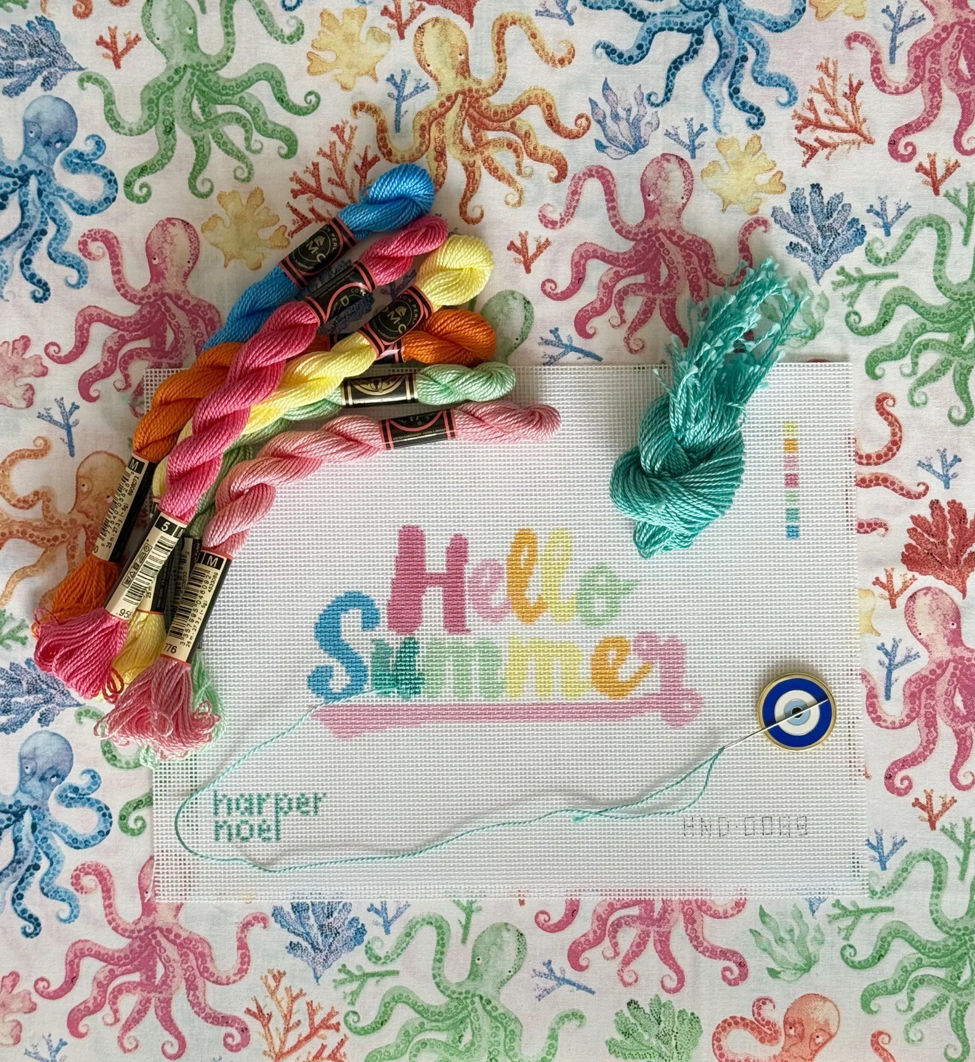 A fabric with a colorful octopus and coral print, along with various embroidery threads and a needle on top of it, and a partially visible embroidered word in rainbow colors that says "Happen".