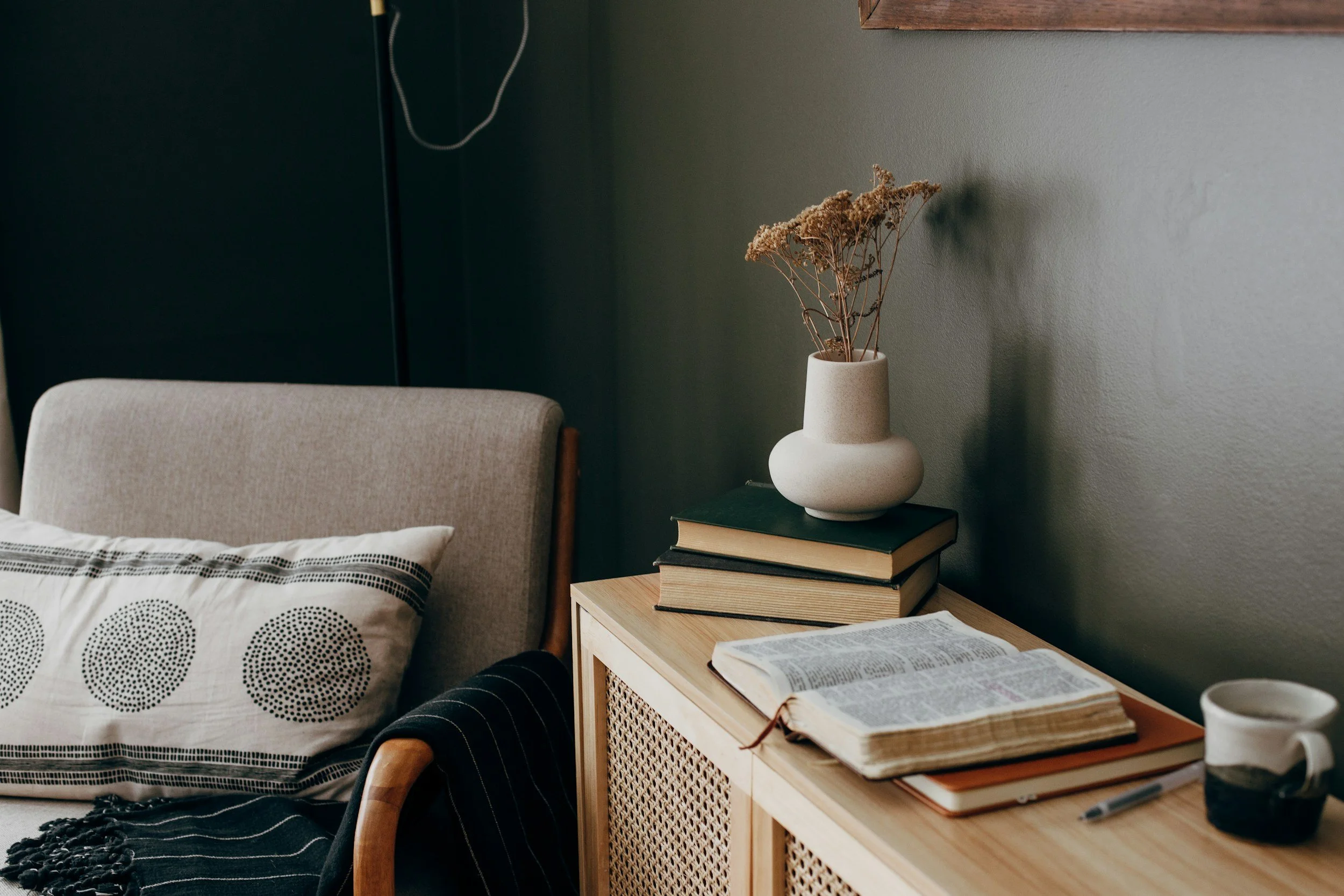 A cozy bedroom corner with a bed that has a white pillow with black dotted circles, a black striped blanket, and a beige upholstered headboard. To the right, a nightstand holds a stack of books, an open book, a ceramic mug, and a white vase with dried flowers. The wall behind is painted in a muted green shade, and a wooden frame is partially visible at the top.