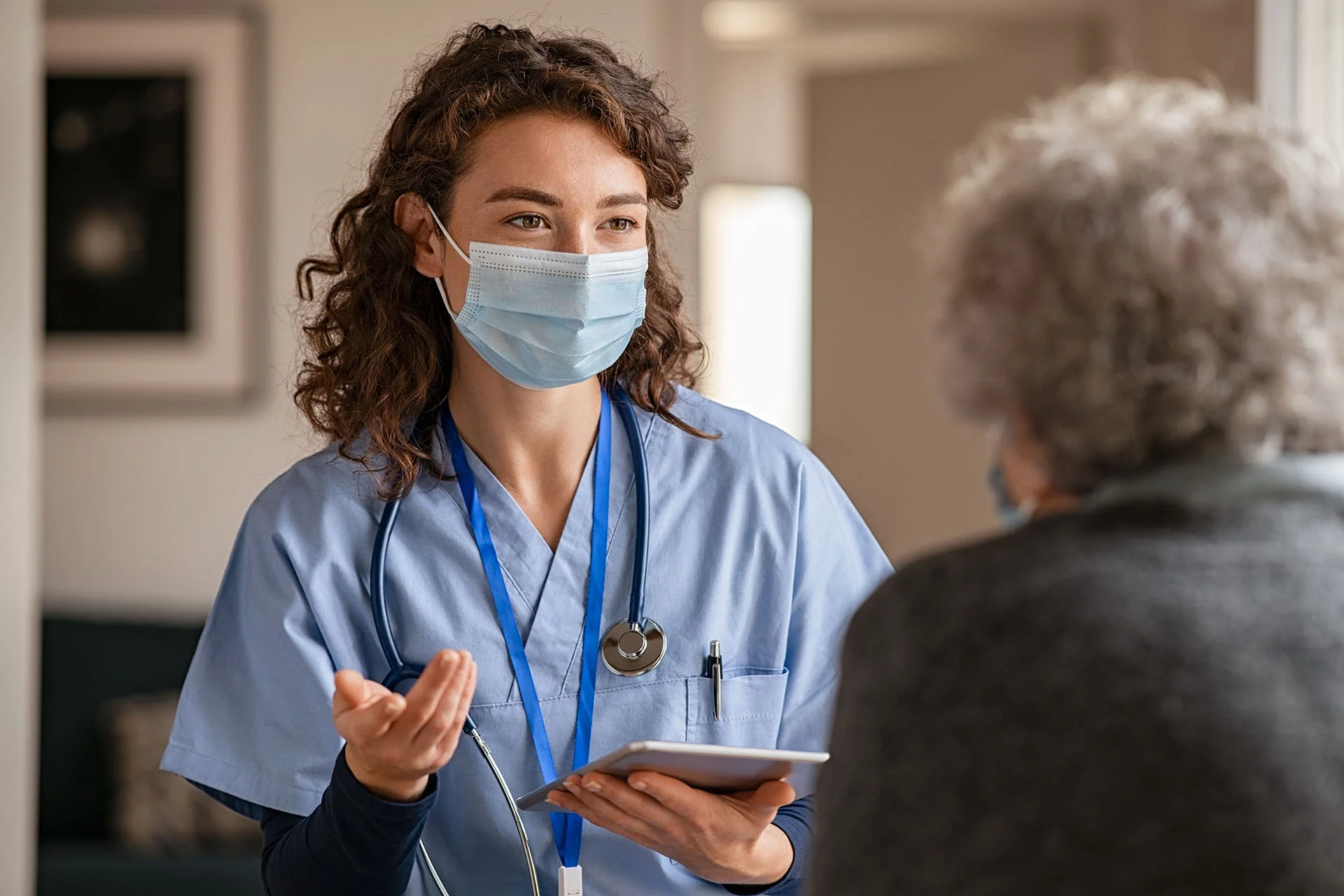 A healthcare worker in a blue scrubs and mask talking to an elderly patient.