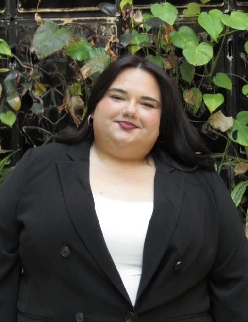 Woman with long dark hair wearing a black blazer and white top standing in front of green leafy plants.