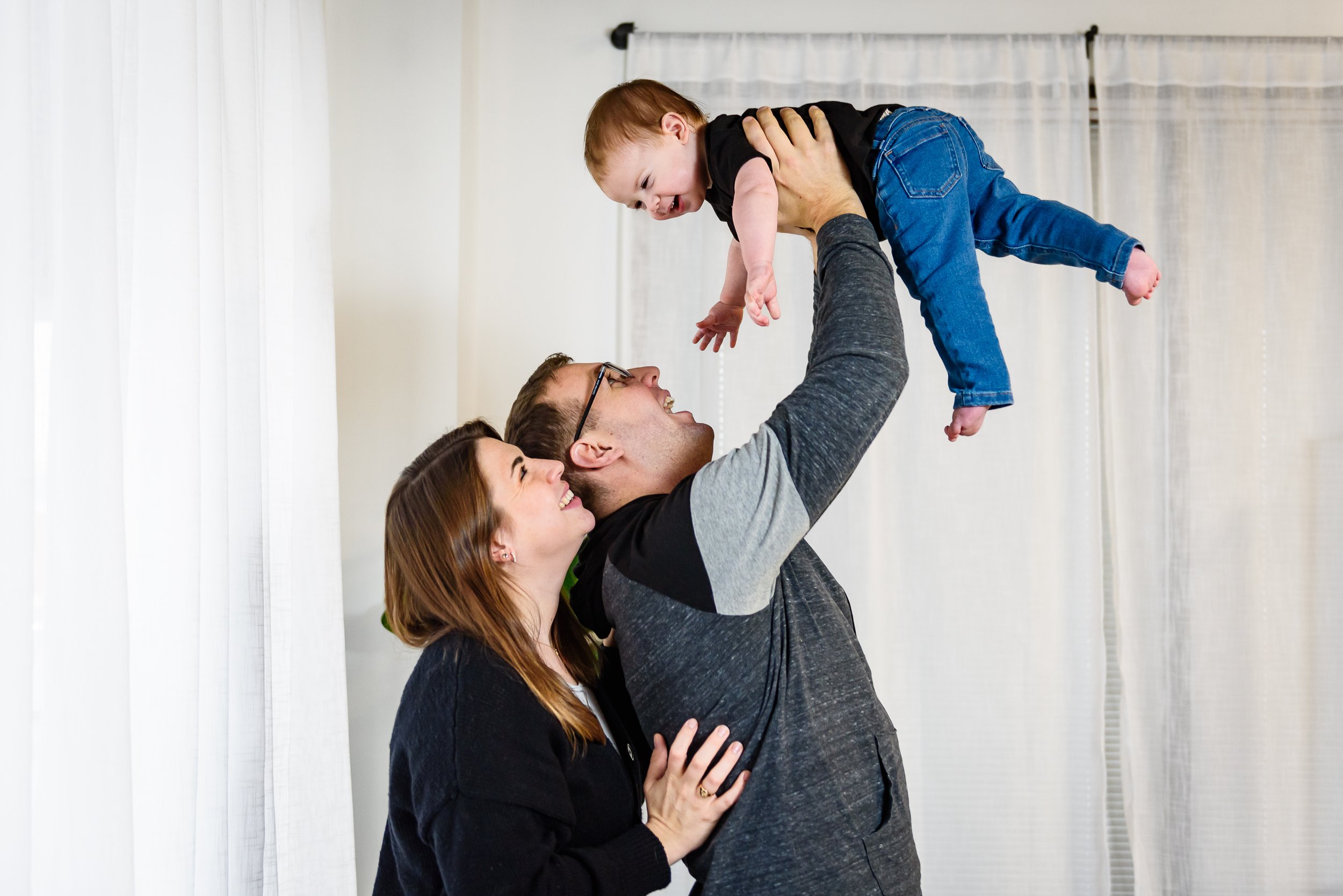 A family of three, mother, father, and young son, playing together indoors. The father lifts the son into the air while everyone smiles and looks at each other.