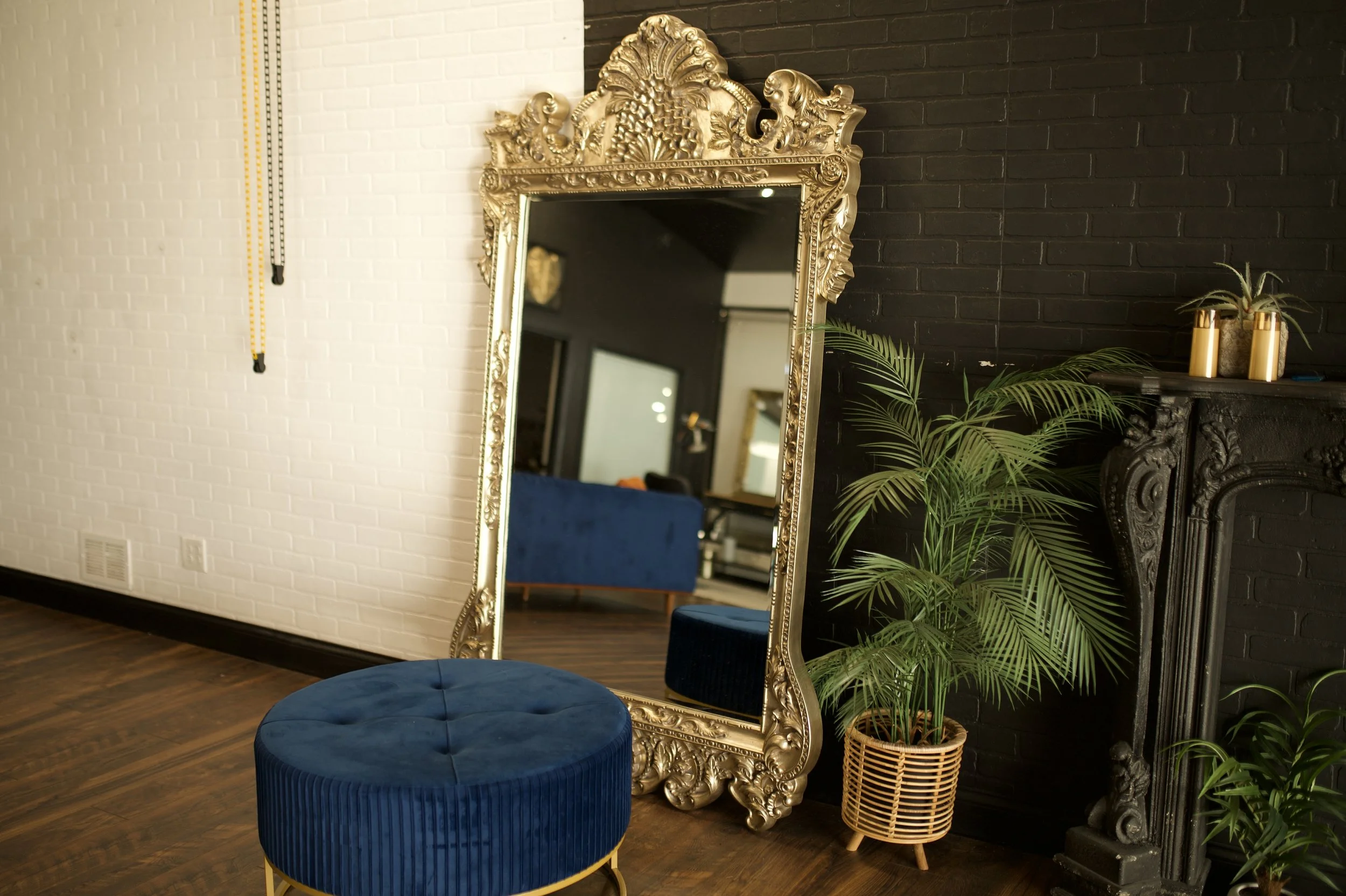An ornate gold-framed full-length mirror leaning against a wall, with a blue velvet ottoman and a potted palm plant nearby, in a room with dark and white brick walls.