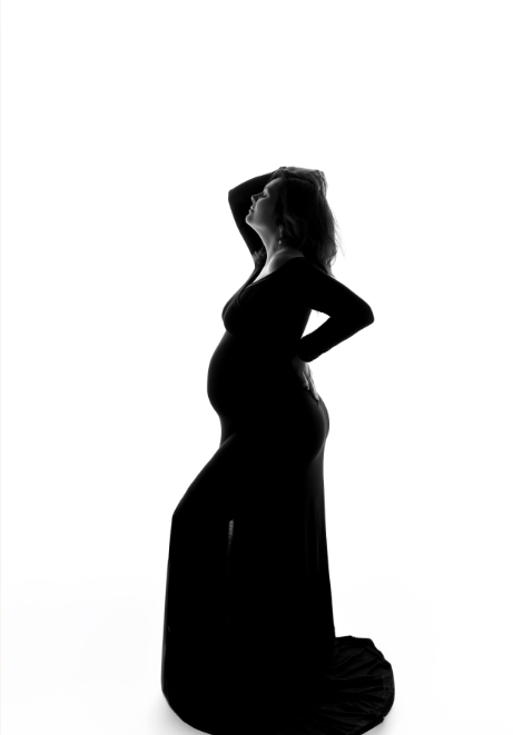 Silhouette of a pregnant woman in a long dress, standing with one hand on her hip and the other behind her head, against a white background.
