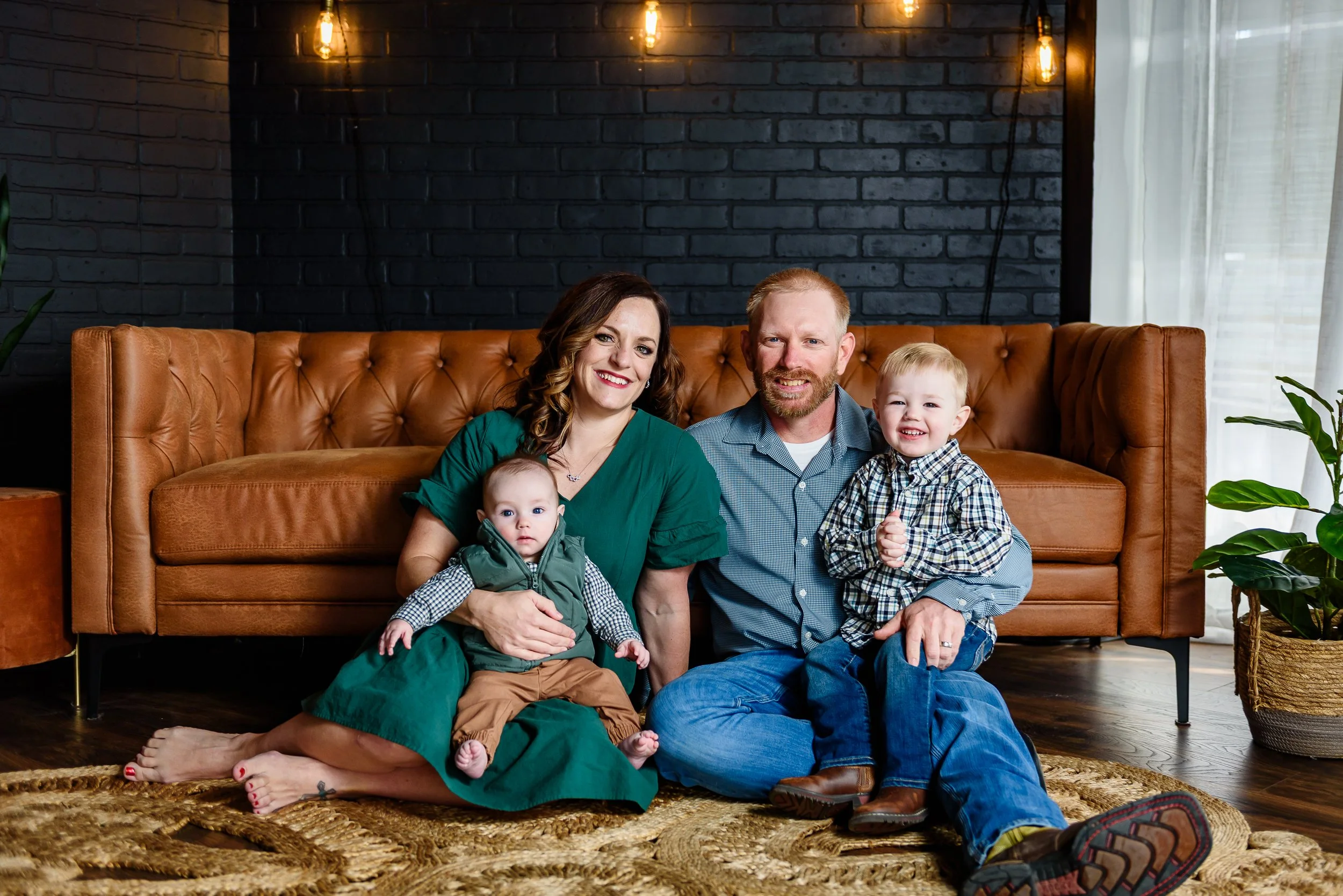 Happy family of four sitting on the floor in front of a brown leather couch, with a black brick wall background and warm lighting, including a mother, father, and two young children, all smiling.