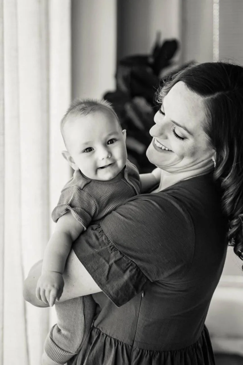 A woman holding a smiling baby in her arms indoors near a window with curtains.