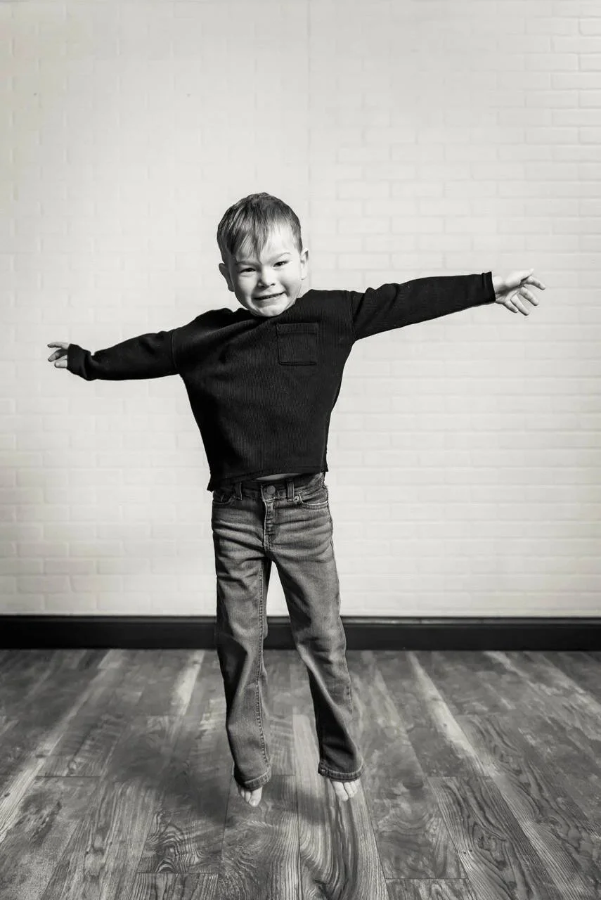 A young boy with short hair jumping in mid-air with arms outstretched, wearing a long-sleeve dark shirt and jeans, against a plain white brick wall background.
