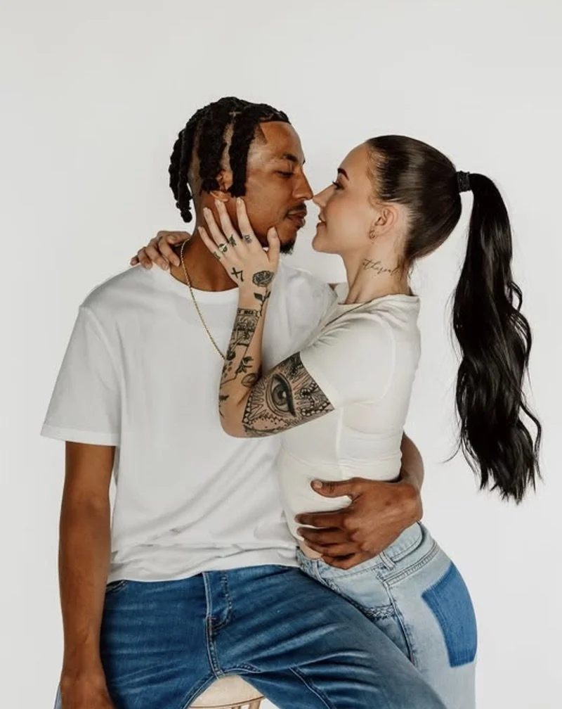 A couple standing close with faces almost touching, the woman holding the man's face, both smiling. They are wearing casual white shirts and jeans, with tattoos visible on both of them, against a plain white background.