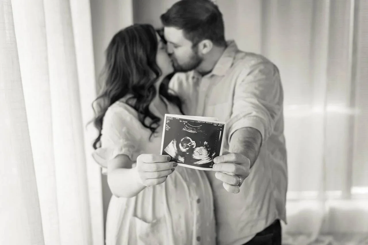 A woman and a man sharing a kiss, holding an ultrasound photo, in a room with light-colored curtains.