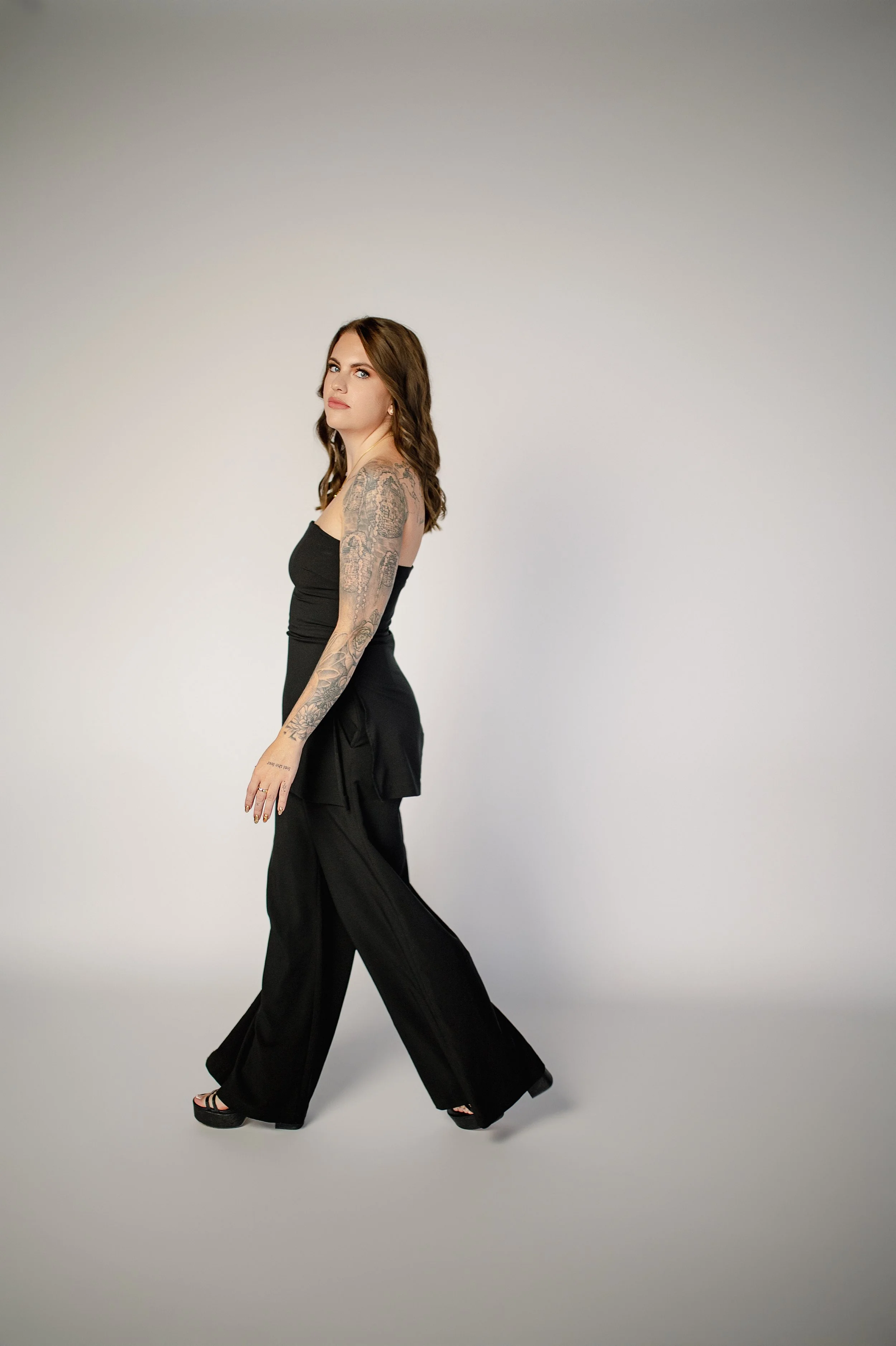 A woman in black wide-leg pants and a strapless top, with tattoos on her left arm, walking in a studio with a plain gray background.