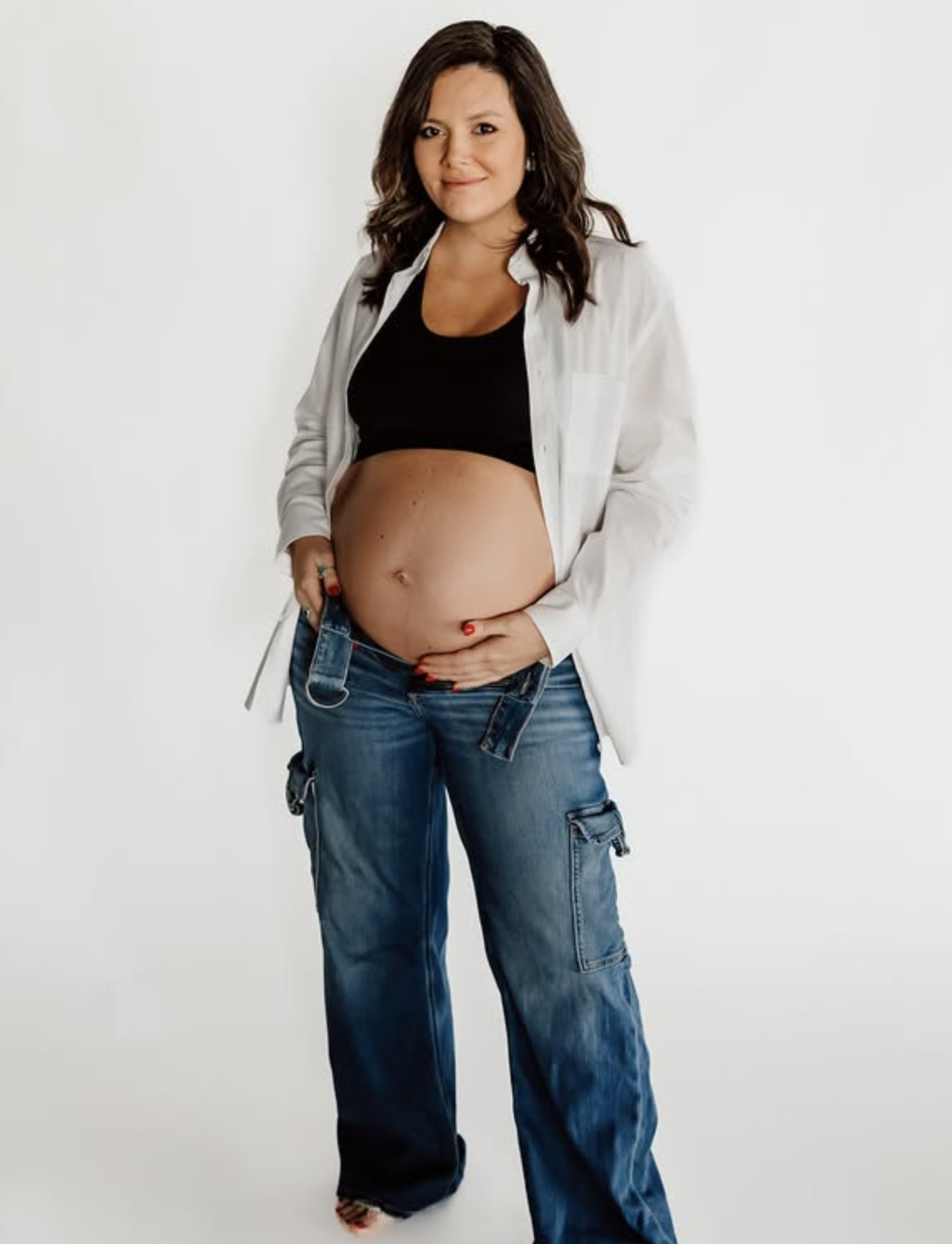 A pregnant woman standing against a plain white background, wearing a black crop top, unbuttoned white shirt, and blue cargo jeans, with one hand resting on her belly.