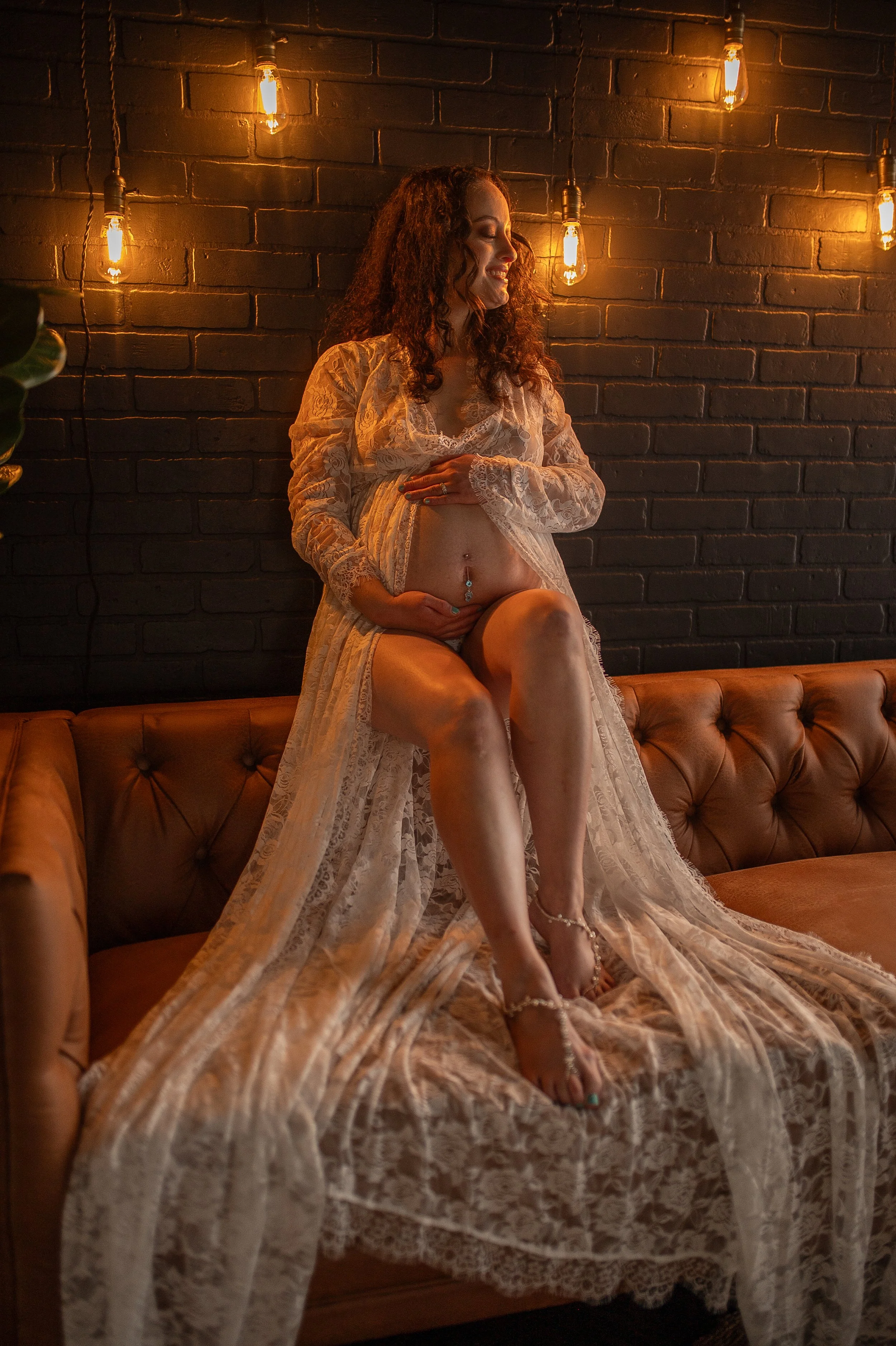 A pregnant woman sitting on a leather couch with a black brick wall behind her. She is wearing a sheer, lace robe and high heels, with her hand on her belly. There are warm hanging light bulbs illuminating the scene.